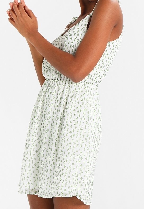 Femme portant une robe blanche sans manches avec un petit imprimé cactus vert, debout les mains jointes près de la poitrine.