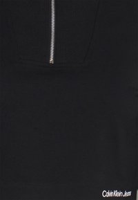 Svart sweatshirt med en zip-försedd ficka och ribbad textur på bröstområdet. "Calvin Klein Jeans"-logotyp i vitt på nedre högra hörnet.