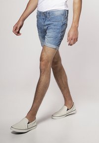 Shorts de mezclilla en azul claro, con un acabado desgastado y puños enrollados, acompañados de zapatos slip-on blancos con detalles en negro.