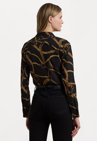 Camicia nera con bottoni, decorata con un motivo a catena e corda dorata. Maniche lunghe, vestibilità sagomata e colletto classico, abbinata a jeans neri.