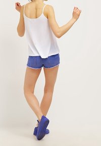 Persona vestida con una camiseta sin mangas blanca, pantalones cortos de mezclilla azules y zapatos azules, de pie con la espalda hacia adelante y los brazos levantados ligeramente.