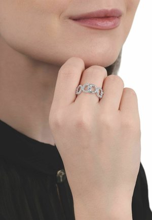 Mano de mujer cerca del rostro que lleva un anillo de plata con formas ovaladas entrelazadas adornadas con piedras preciosas claras.