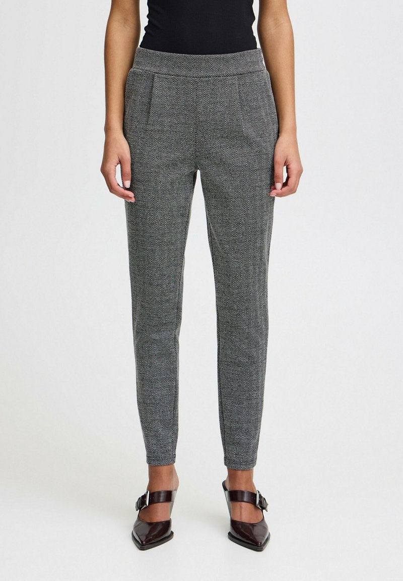 Femme portant un pantalon gris ajusté à motif chevrons avec taille élastique et des chaussures noires à bout pointu avec brides à la cheville.