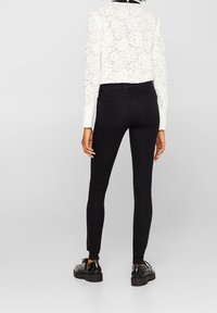 Jeans negros ajustados con cintura alta, forma ceñida y diseño clásico de cinco bolsillos, combinados con una blusa blanca de encaje de manga larga y zapatos negros.