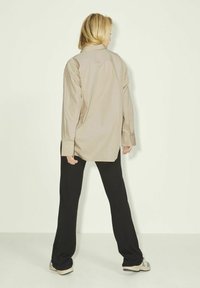Camicetta beige a maniche lunghe con colletto classico, taglio dritto e polsini con bottoni, abbinata a pantaloni neri a gamba larga e sneakers grigie.