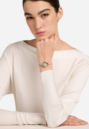 Silverfärgad armbandsur med rund urtavla, grå läderrem och minimalistisk design, bärs av en person i en slät, ljus långärmad topp.