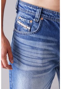 Niebieskie jeansy z denimu o klasycznym kroju, wyblakłym wyglądzie, miedzianych nitach i małą kieszonką z materiałowym akcentem. Widoczne szczegóły tekstury powierzchni.