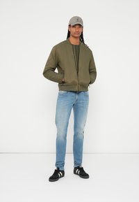 Oliven grøn quiltet bomberjakke med lynlås foran, parret med slidte blå jeans og sorte sneakers med hvide striber. Grå kasket på hovedet.