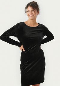 Robe en velours noir avec des manches longues, un col rond et une silhouette ajustée qui tombe doucement. Texturé lisse avec un léger brillant.