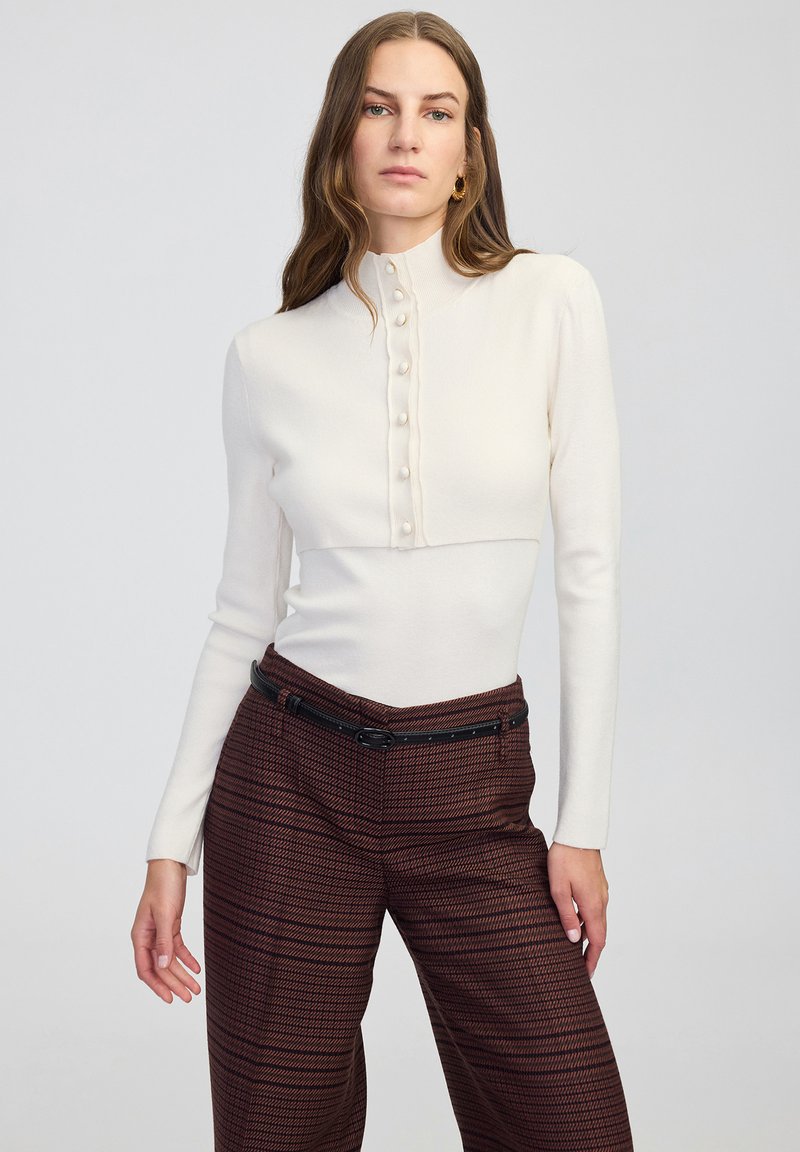 Suéter blanco ajustado con un cuello alto y cierre frontal de botones. Combinado con pantalones de pierna ancha de patrón marrón y una cintura ceñida con cinturón negro.