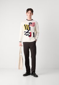 Pull en tricot crème avec des motifs multicolores de drapeaux et à carreaux. Associé à un pantalon foncé et des chaussures noires, portant un sac fourre-tout beige.