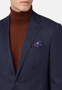 Blazer navy con un sutil patrón de cuadros, jersey de cuello alto marrón debajo y un pañuelo de bolsillo en paisley en tonos de azul, rojo y blanco.