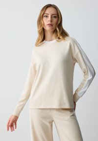 Top lungo maniche color beige chiaro con scollo rotondo e finiture laterali bianche. Tessuto morbido, vestibilità comoda e polsini a costine. Abbinato a pantaloni coordinati.