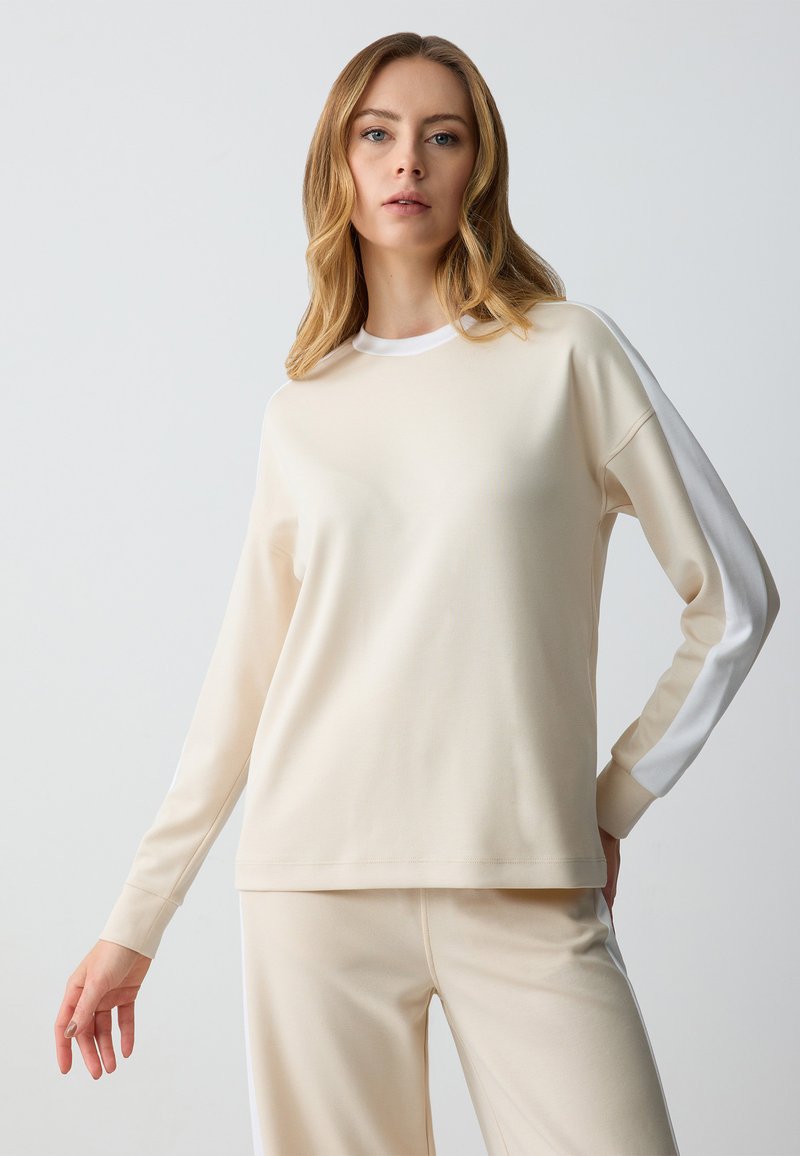 Top lungo maniche color beige chiaro con scollo rotondo e finiture laterali bianche. Tessuto morbido, vestibilità comoda e polsini a costine. Abbinato a pantaloni coordinati.