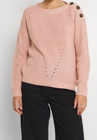 Pull rose clair en maille avec un motif texturé, des poignets côtelés et trois boutons marron sur l'épaule. À porter avec un pantalon noir.