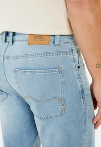 Jasnoniebieskie dżinsy z denimu z brązową skórzaną naszywką z napisem "camel active". Szczególne przeszycia i standardowe tylne kieszenie. Gładka tekstura.