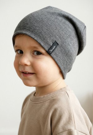 Kleinkind lächelt seitlich, trägt eine graue Strickmütze und ein beiges Hemd vor einem einfarbigen hellen Hintergrund.