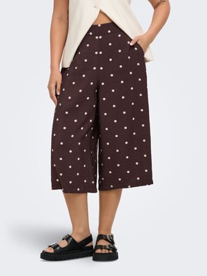 Femme portant un pantalon marron large et court avec des pois blancs, associé à des sandales noires à boucle et un haut blanc sans manches.