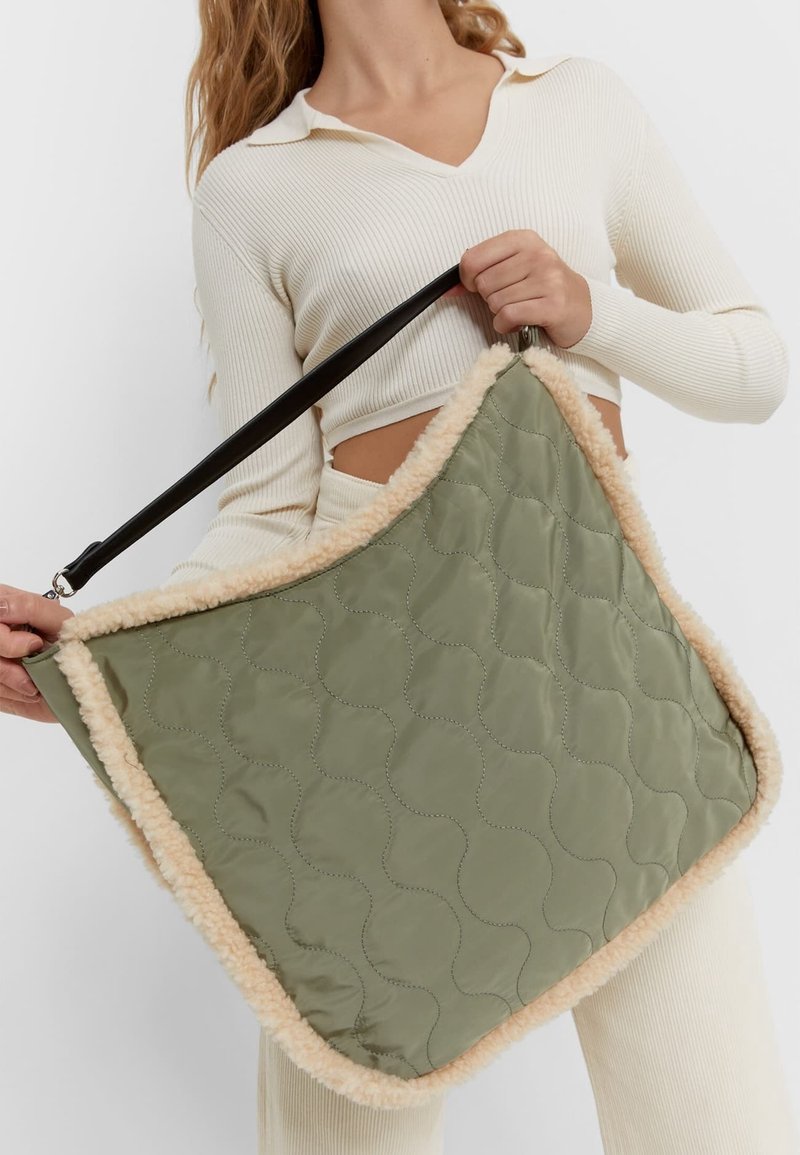 Personne en tenue côtelée crème tenant un grand sac matelassé vert olive avec une bordure en shearling beige et une sangle noire.