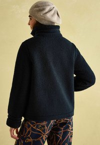 Čierna fleecová bunda s vysokým golierom a textúrovanou tkaninou. Dizajn obsahuje široké rukávy a voľný strih, kombinovaný so vzorovanými nohavicami.