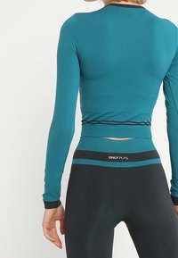 Donna che indossa un crop top a maniche lunghe teal e leggings neri a vita alta con strisce teal e logo "ONLY PLAY" in vita.