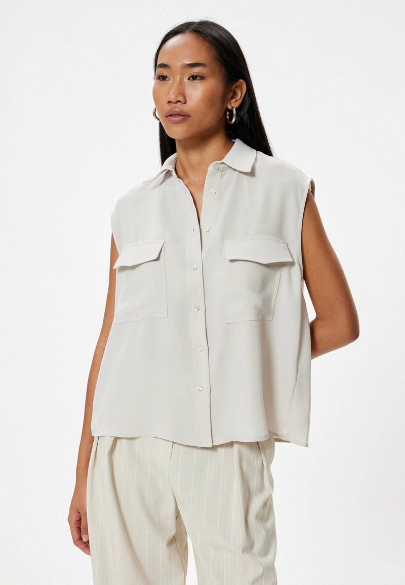 Helle beige ärmellose Bluse mit Kragen, ausgestattet mit zwei Fronttaschen und Knopfverschluss. Glattes Material mit lässiger Passform.