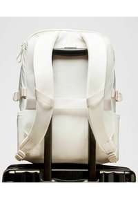 Mochila de tela blanca con correas ajustables, que presenta una textura suave y un bolsillo. Colocada sobre el asa de una maleta negra.