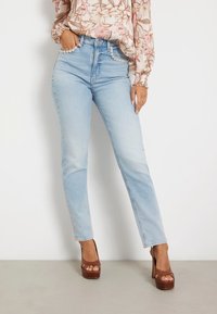 Mujer vestida con unos pantalones vaqueros de tiro alto en azul claro, adornados con perlas en los bolsillos, blusa de flores de manga larga y tacones de plataforma marrones.