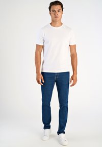 Biała bawełniana koszulka z okrągłym dekoltem, zestawiona z niebieskimi dżinsami. Model nosi białe trampki, stojąc na tle jednolitego tła.