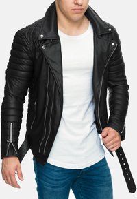 Giacca da motociclista in pelle nera con maniche trapuntate, cerniere argentate e dettaglio in vita con cintura. Indossata sopra una t-shirt bianca.