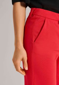 Rode op maat gemaakte broek met voorplooien, gemaakt van gladde stof, met riemlussen en een aansluitende snit. Close-up van een hand die op de zijkant rust.