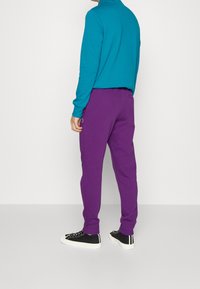 Türkisfarbene Langarmbluse kombiniert mit lila Jogginghosen. Die Hose hat eine lockere Passform und elastische Bündchen, ergänzt durch schwarze Turnschuhe.