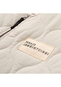 Quilted cream jacket with a zipper closure. Features a fabric label with "HALO" and numbers, displayed on a textured surface.