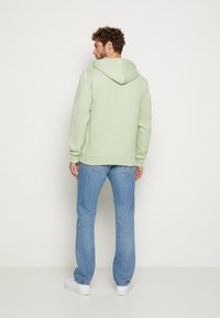 Mintgroene hoodie met een kangoeroezak en een trekkoord in de capuchon, gecombineerd met lichtblauwe jeans en witte sneakers, gezien vanuit de achterkant.