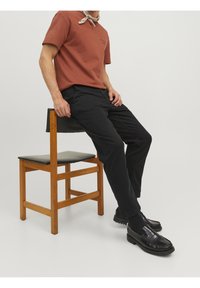 T-shirt di cotone marrone con un piccolo logo, abbinato a pantaloni neri e mocassini neri lucidi. La sedia presenta una struttura in legno e seduta nera.