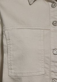 Ljusbeige denimtyg med en ficka och silverfärgade tryckknappar. Har en klassisk krage och synliga sömnadsdetaljer.
