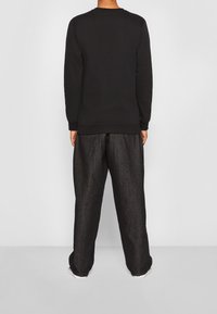 Persona de pie de espaldas a la cámara, vistiendo una camiseta negra de manga larga y pantalones negros holgados, contra un fondo claro y liso.