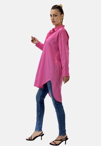 Langes pinkes Hemd mit Kragen, seitlichen Schlitzen und geknöpften Manschetten, kombiniert mit blauen Skinny-Jeans und schwarzen Sandalen mit Absatz. Glatte Stoffstruktur.