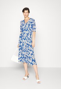 Vestido floral azul com padrões de flores brancas, mangas curtas e volumosas, decote em V e comprimento médio; combinado com sandálias brancas de salto bloco.