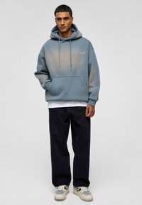 Capuchon sweatshirt in vervaagde blauw, met een voorzak en koordjes. Gecombineerd met donkere spijkerbroeken en lichtgekleurde sneakers.