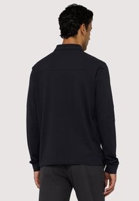 Tmavě modrá košile s dlouhým rukávem, měkkou texturou, límcem a postranními rozparky na lemu. Má kontrastní prošívání podél ramen.