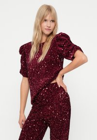 Blonde vrouw poseert in een bijpassende bordeauxrode fluwelen top met pailletten en pofmouwen, samen met een broek, tegen een effen witte achtergrond.