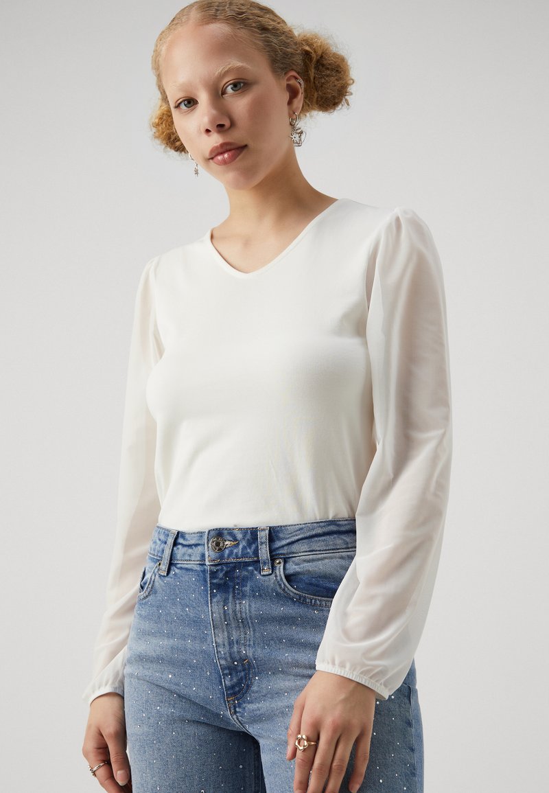 Witte V-hals top met doorschijnende, geplooide lange mouwen, gecombineerd met hooggewonden blauwe denim jeans met parelversieringen.