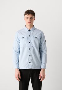 Hele sinine nööbitav shirts, millel on kaks rinna taskut, mustad nupud ja pikad varrukad. Valmistatud siledast kangast klassikalise kraega.