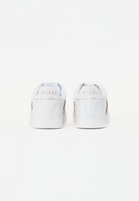 Vita sneakers med en slät läderfinish, gyllene "GUESS"-logotyp baktill och en kontrasterande beige accent på sidorna.