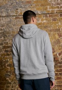 Hellegrauer Fleece-Hoodie mit Kordelzugkapuze, gerippten Bündchen und Saum, von hinten gegen eine strukturierte Ziegelwand gezeigt.