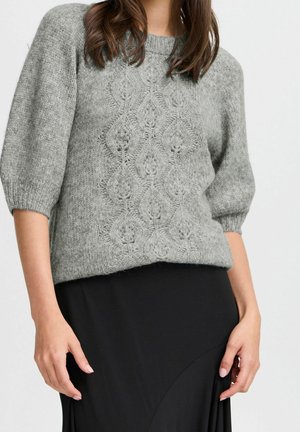 Woman wearing a grey knitted three-quarter sleeve sweater with a textured pattern and a black skirt, standing with hands relaxed.