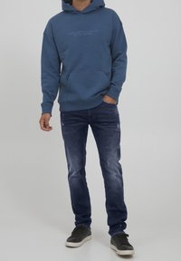 Blue hoodie with front pocket and text logo, paired with dark denim jeans featuring slight distressing, and black sneakers.