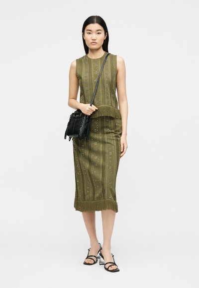 Γυναίκα που φορά ένα αμάνικο μίντι φόρεμα σε ελιά πράσινο με σχέδια και κρόσσια, μαύρη τσάντα χιαστί και μαύρα ψηλοτάκουνα σανδάλια, στέκεται μπροστά σε λευκό φόντο.