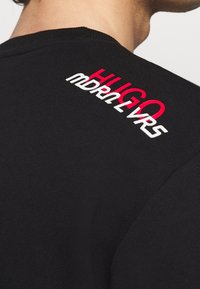 Camiseta negra de algodón con un cuello acanalado. Texto bordado en rojo y blanco "HUGO" y "mdrn2vrs" en el hombro trasero.